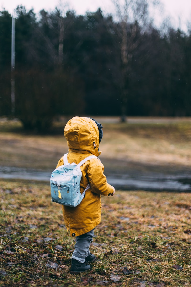 A small child in a yellow coat walking by a river, photographed from behind